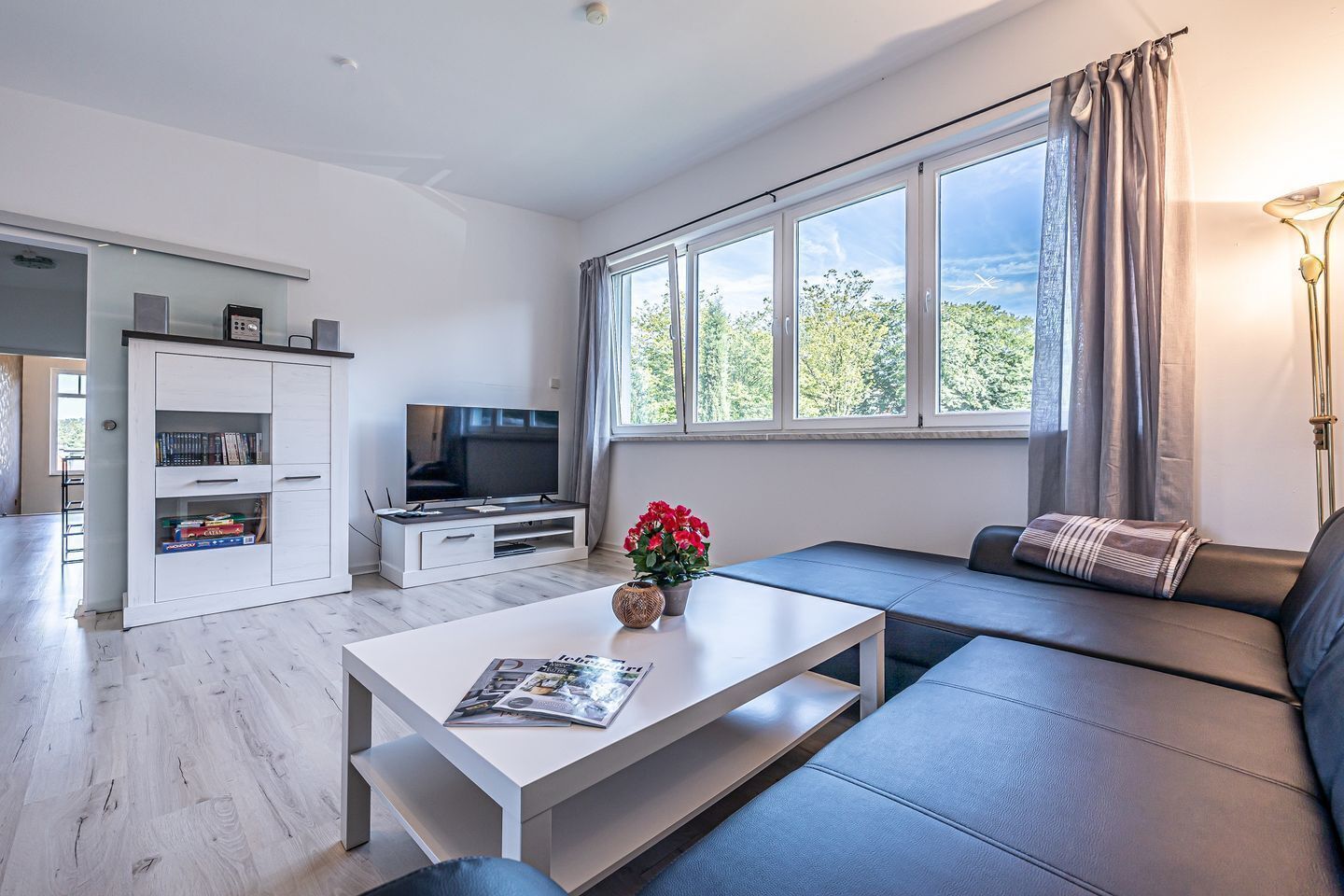 Ein modernes Wohnzimmer mit einem Flachbildfernseher, einem weißen Couchtisch mit einer Zeitschrift, einem Sofa und einem großen Fenster mit Vorhängen.