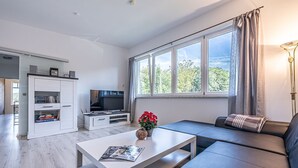 Ein modernes Wohnzimmer mit einem Flachbildfernseher, einem weißen Couchtisch mit einer Zeitschrift, einem Sofa und einem großen Fenster mit Vorhängen.