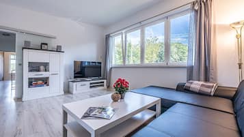 Ein modernes Wohnzimmer mit einem Flachbildfernseher, einem weißen Couchtisch mit einer Zeitschrift, einem Sofa und einem großen Fenster mit Vorhängen.