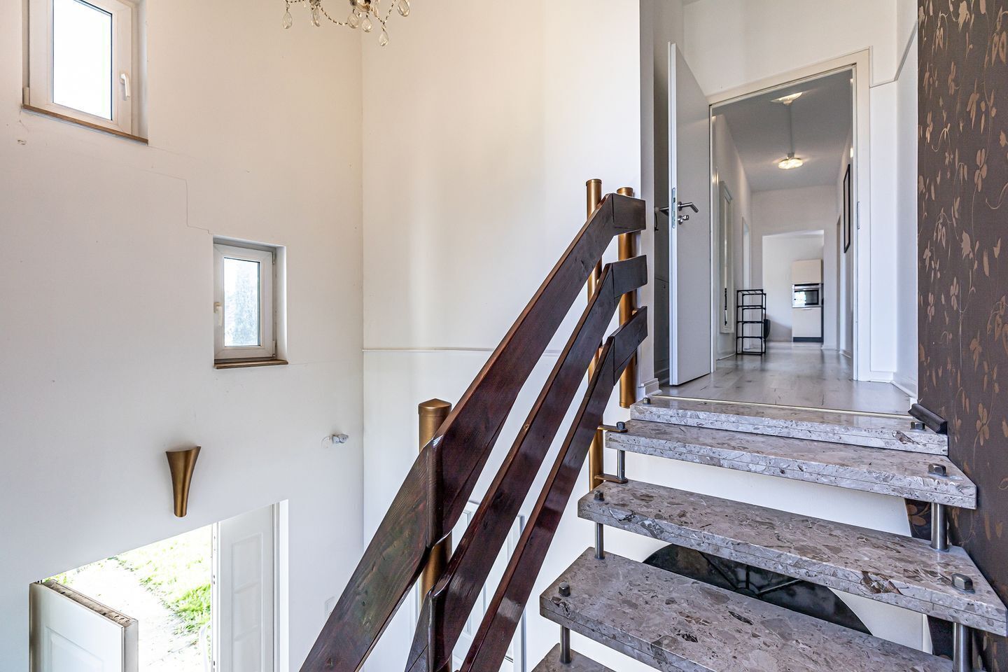 Eine Treppe mit Holzboden und Metallgeländern, die zu einem Flur führt, von dem aus man eine Küche und einen Essbereich sieht.