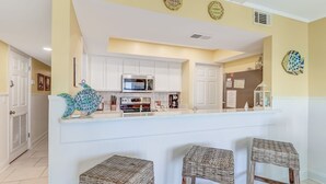 A kitchen with white cabinets, a microwave, a coffee maker, and a decorative fish wall art. There are three wicker stools in front of the counter. - Beachfront Oasis on Amelia Island Condo - 2 Bedroom Ketch Courtyard (Fernandina Beach)