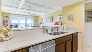 A kitchen with a white countertop, stainless steel sink, and wooden cabinets. There are framed pictures on the wall and a window with a view of the ocean. - Beachfront Oasis on Amelia Island Condo - 2 Bedroom Ketch Courtyard (Fernandina Beach)