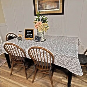 Una mesa de comedor con mantel estampado, dos sillas de madera, un florero con flores y un cartel de bienvenida.