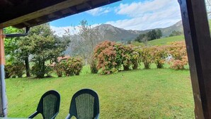 A covered outdoor seating area with two chairs, a grassy lawn, and a view of mountains and shrubs.