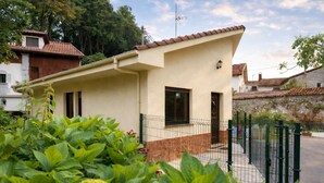 A small, single-story building with a tiled roof, a fenced yard, and lush greenery in the foreground. - Casa la Aldea de Barro (Barro (Llanes))
