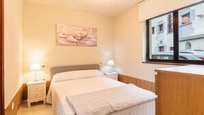 A neatly arranged bedroom with a bed, bedside lamps, a nightstand, a wooden headboard, a window with a view of buildings, and a wall-mounted artwork. - Casa la Aldea de Barro (Barro (Llanes))