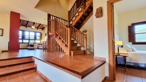 Amplia sala de estar con una escalera de madera, un ventanal con persianas y un rincón de descanso con un sofá y sillones.