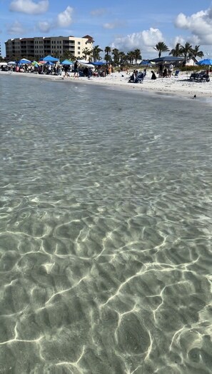 A beach with clear shallow waters, palm trees, and a hotel in the background.