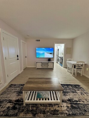 Un salon équipé d’un téléviseur à écran plat, un coin repas avec une table et des chaises, et une cuisine visible à travers une porte ouverte.