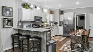 Uma cozinha moderna com armários brancos, uma ilha central com banquetas, eletrodomésticos de aço inoxidável e iluminação pendente.