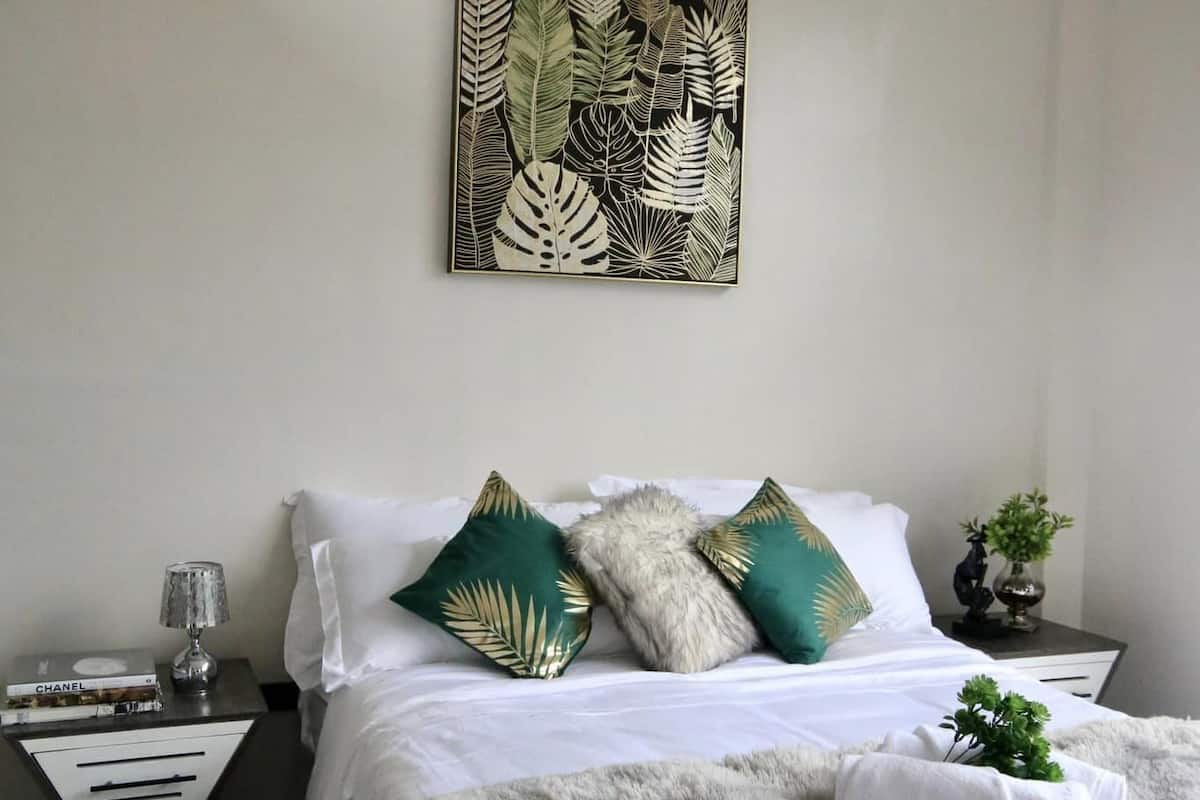 Ein ordentlich eingerichtetes Schlafzimmer mit einem Bett, Kissen, einer Wanddekoration, einem Nachttisch mit Lampe und einer kleinen Pflanze.