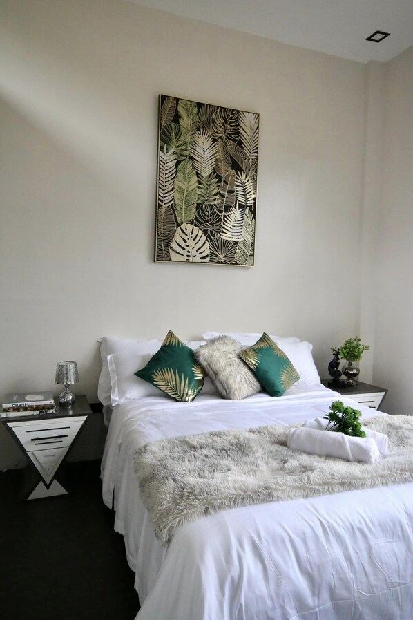 A neatly arranged bedroom with a bed, pillows, a decorative wall piece, a nightstand with a lamp, and a small plant. - Fight Club Philippines (Puerto Princesa)