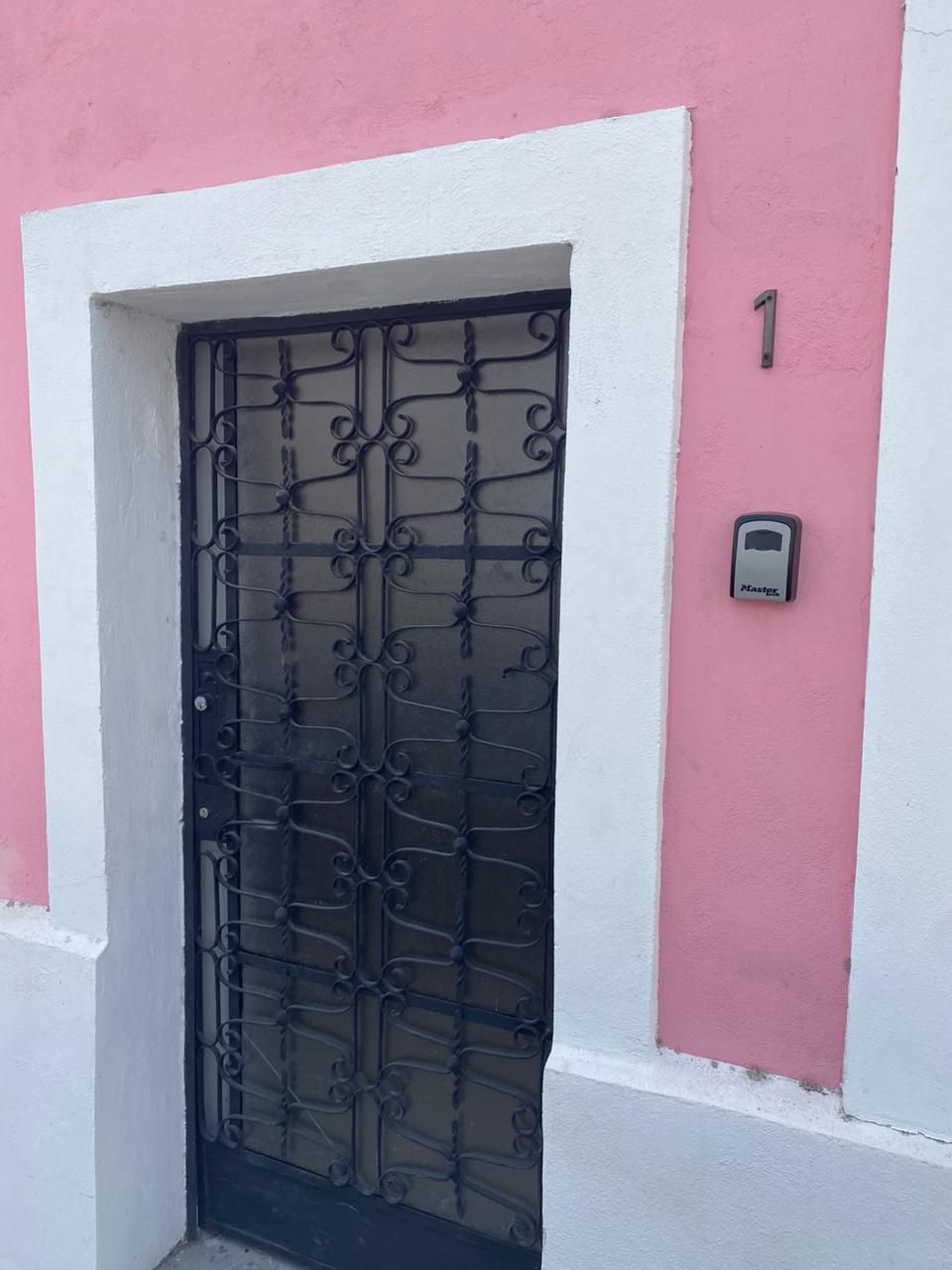 Una puerta con reja de hierro forjado, un marco blanco y una pared rosa con el número '1' encima.