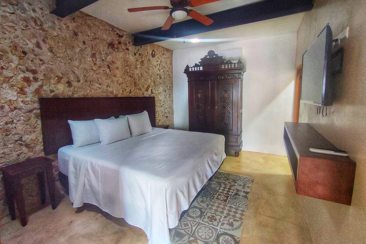 Une chambre avec un mur en pierre, un lit recouvert de draps blancs, une tête de lit en bois, un ventilateur de plafond, une armoire en bois et un téléviseur fixé au mur.