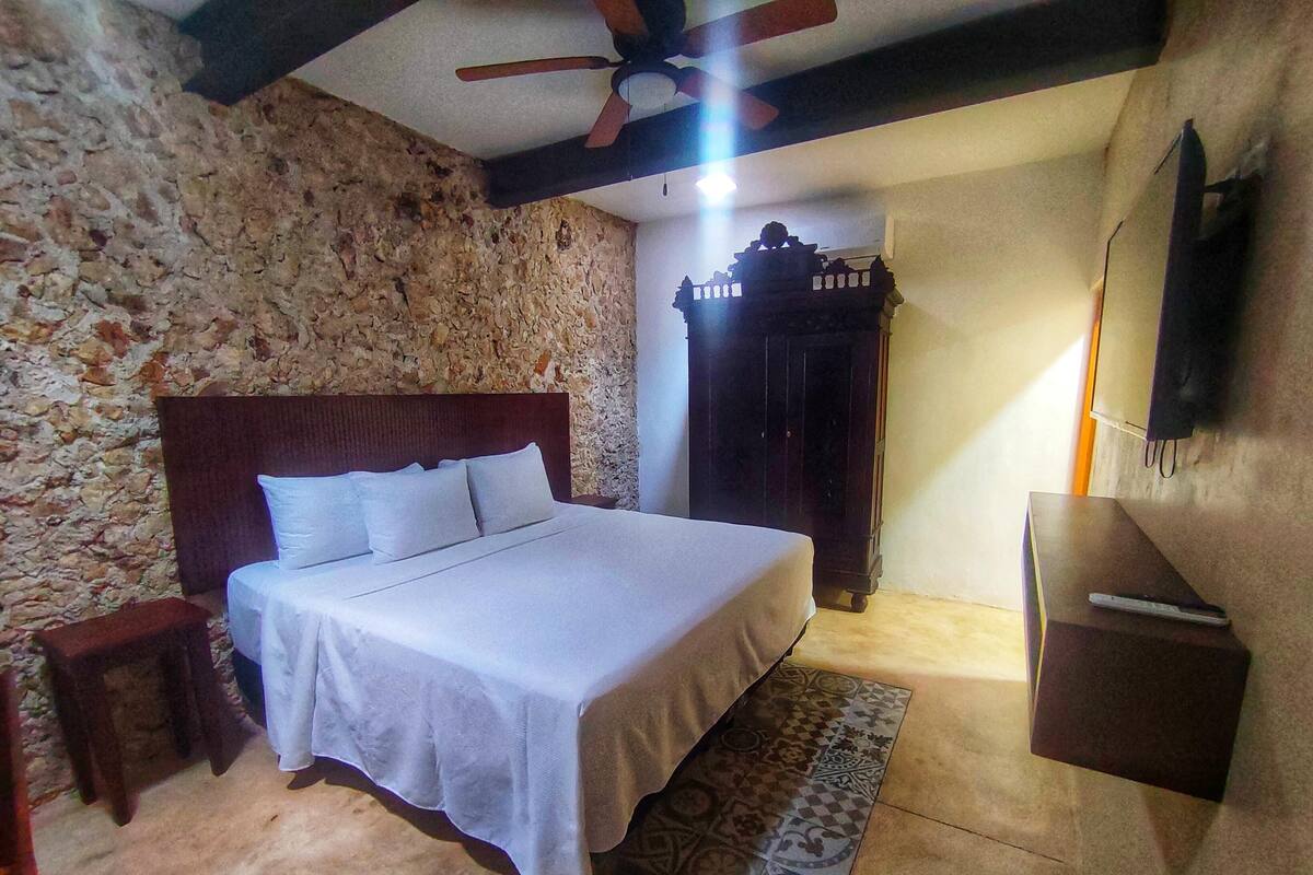 Une chambre avec un mur en pierre, un lit en bois, une armoire en bois foncé et un ventilateur de plafond.