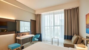 Ein modernes Hotelzimmer mit einem großen Bett, einem Schreibtisch mit Stuhl, einer kleinen Couch, einem Fernseher und einem großen Fenster mit Vorhängen.