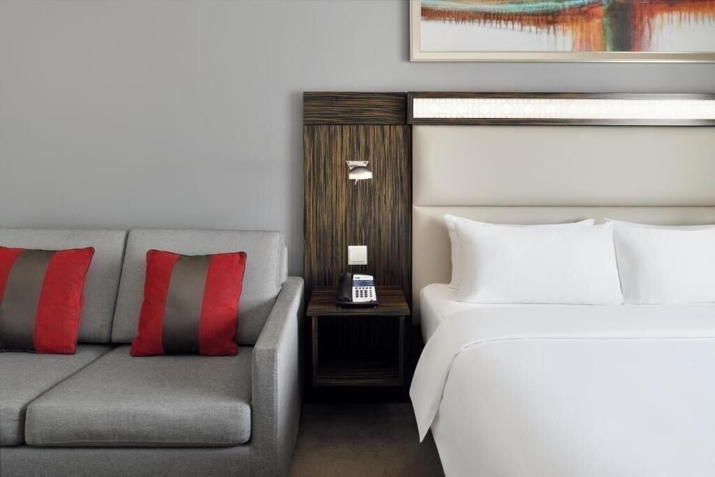 Une chambre d’hôtel avec un canapé gris, une table de chevet avec une lampe et un lit avec des oreillers blancs.