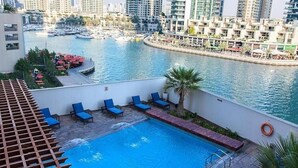 A rooftop pool with blue lounge chairs, a palm tree, and a marina with boats in the background.