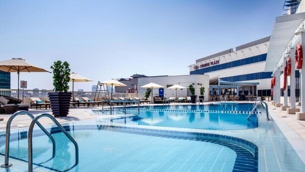 A rooftop swimming pool with lounge chairs and umbrellas, surrounded by a modern building with a blue and white facade.