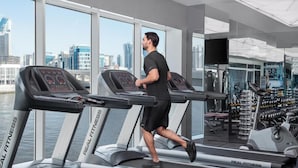 A man running on a treadmill in a modern gym with large windows and a city view. - Standard Room In Business Bay By E R (Dubai)
