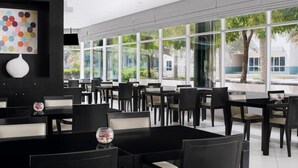 A modern dining area with black tables and chairs, large windows, and a colorful abstract art piece on the wall. - Standard Room In Media City (Dubai)