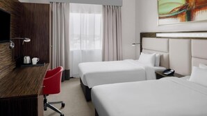 A hotel room with two beds, a desk, a red chair, a coffee maker, and a painting on the wall. - Standard Room In Media City (Dubai)