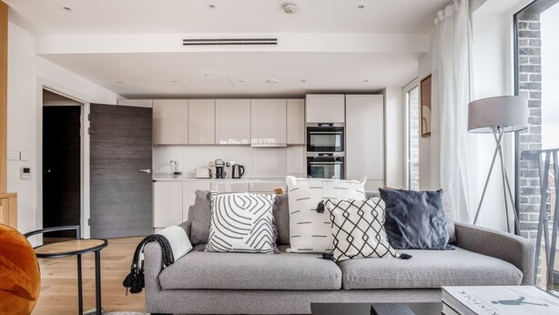 A modern living room with a grey sofa, a coffee table, and a bookshelf. There is a kitchen in the background with white cabinets and a microwave.