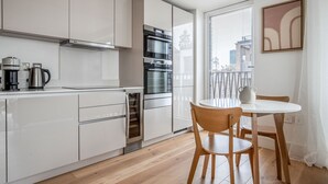 A modern kitchen with white cabinets, a built-in oven, and a coffee maker. There is a dining area with a round table and two chairs.
