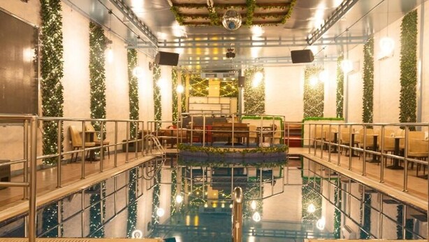 An indoor swimming pool with a central diving platform, surrounded by seating areas and walls adorned with greenery.