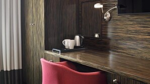 A hotel room with a wooden panel wall, a small desk with a kettle, a cup, and a tray, and a red chair.