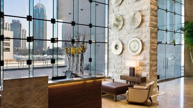 Un espace d’accueil moderne avec un comptoir de réception en marbre, un coin salon avec un canapé et des fauteuils, des éléments décoratifs fixés au mur et une vue sur le paysage urbain grâce à de grandes fenêtres.