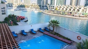 Una piscina en la azotea con sillas reclinables azules, una palmera y un puerto deportivo con barcos al fondo.