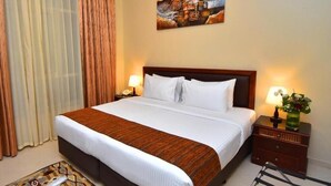 A hotel room with a large bed, a nightstand with a lamp, a patterned rug, and a painting on the wall. - Studio Apartment Near Al Khan Beach By E R (الشارقة)