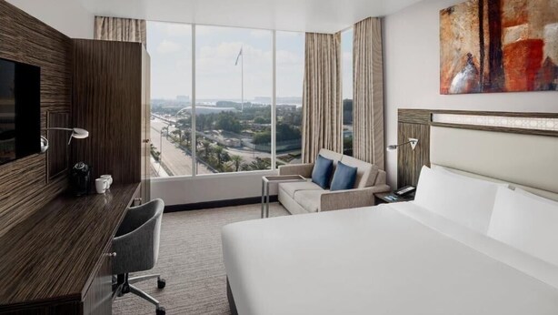 A hotel room with a large bed, a desk, a chair, a sofa, and a large window offering a city view.