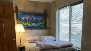 Ein Schlafzimmer mit Bett, einem Gemälde, einer Lampe und einem Fenster mit Jalousien.