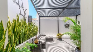 A sunlit outdoor seating area with a pergola, potted plants, and a wall-mounted decorative piece.