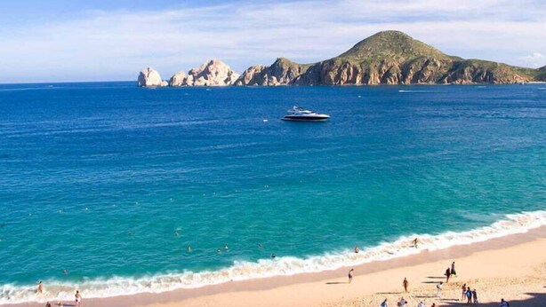 A beach with clear blue water, a boat, and rocky cliffs in the background.
