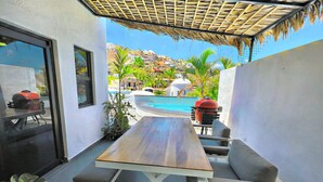 A patio with a wooden table and cushioned seating, a pool, and a view of a hillside with buildings.