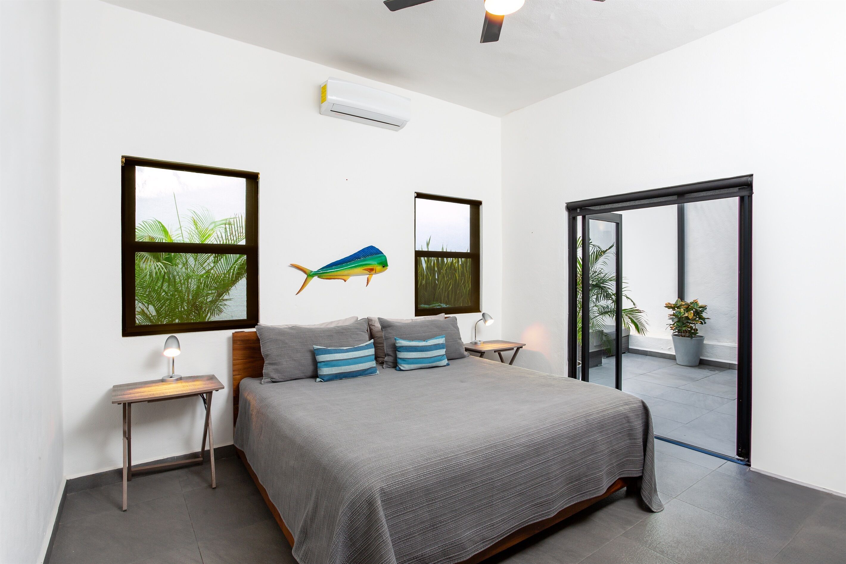 A bedroom with a bed, two nightstands, a wall-mounted air conditioner, and a view of the outdoors through a sliding door.