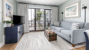 A modern living room with a grey sofa, a flat-screen TV, a wooden coffee table, and a sliding glass door leading to a balcony. - The Shores Lake View Condo Unit 5689 (Williamsburg)