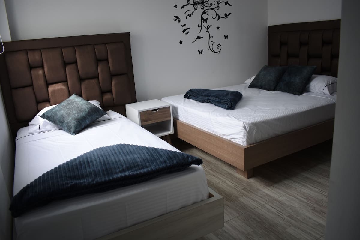 Quarto de hotel com duas camas, cada uma com cabeceira de madeira e roupa de cama branca. Há uma pequena mesa de cabeceira com gaveta e um adesivo de árvore com folhas e estrelas na parede.