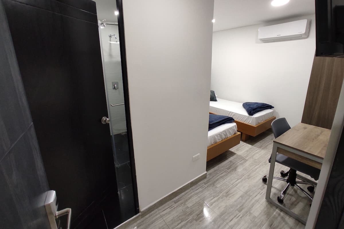 Quarto de hotel moderno com porta escura, banheiro pequeno, duas camas de solteiro, escrivaninha de madeira e uma cadeira.