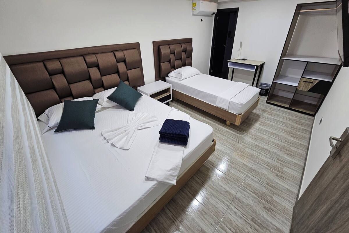 Quarto de hotel com duas camas, um guarda-roupa, uma mesa de cabeceira e uma lâmpada no teto.