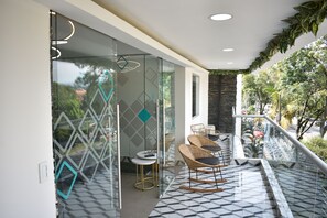 Un interior moderno con divisiones de vidrio, suelo con patrón geométrico, sillas de mimbre y un balcón con vistas a zonas verdes.