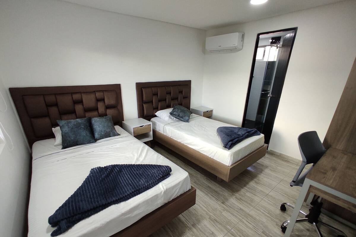 Quarto de hotel com duas camas, uma escrivaninha, uma cadeira e um aparelho de ar condicionado.