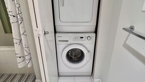 A compact laundry room with a front-loading washer and dryer, a towel rack, and a striped rug on the floor. - Modern 1BR w/ Balcony | 10 Mins Walk to BC Place & Rogers Arena (Vancouver)