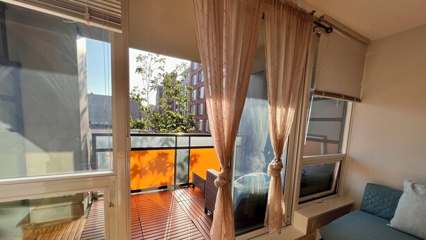 A balcony with a view of buildings and trees, a wooden floor, and a blue sofa with a cushion. - Modern 1BR w/ Balcony | 10 Mins Walk to BC Place & Rogers Arena (Vancouver)
