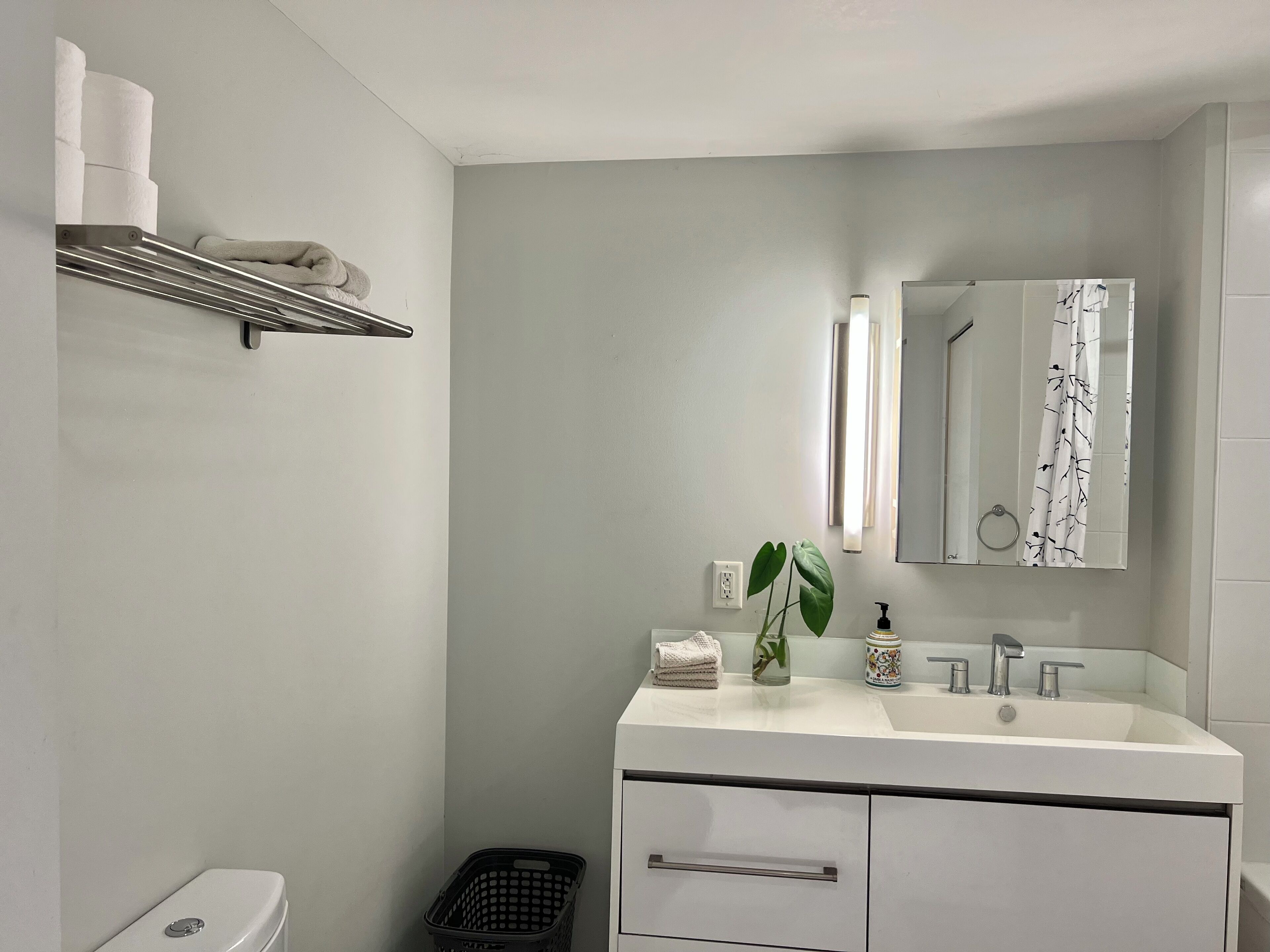 A modern bathroom with a white sink, mirror, and toilet. There is a shelf with towels and a basket.