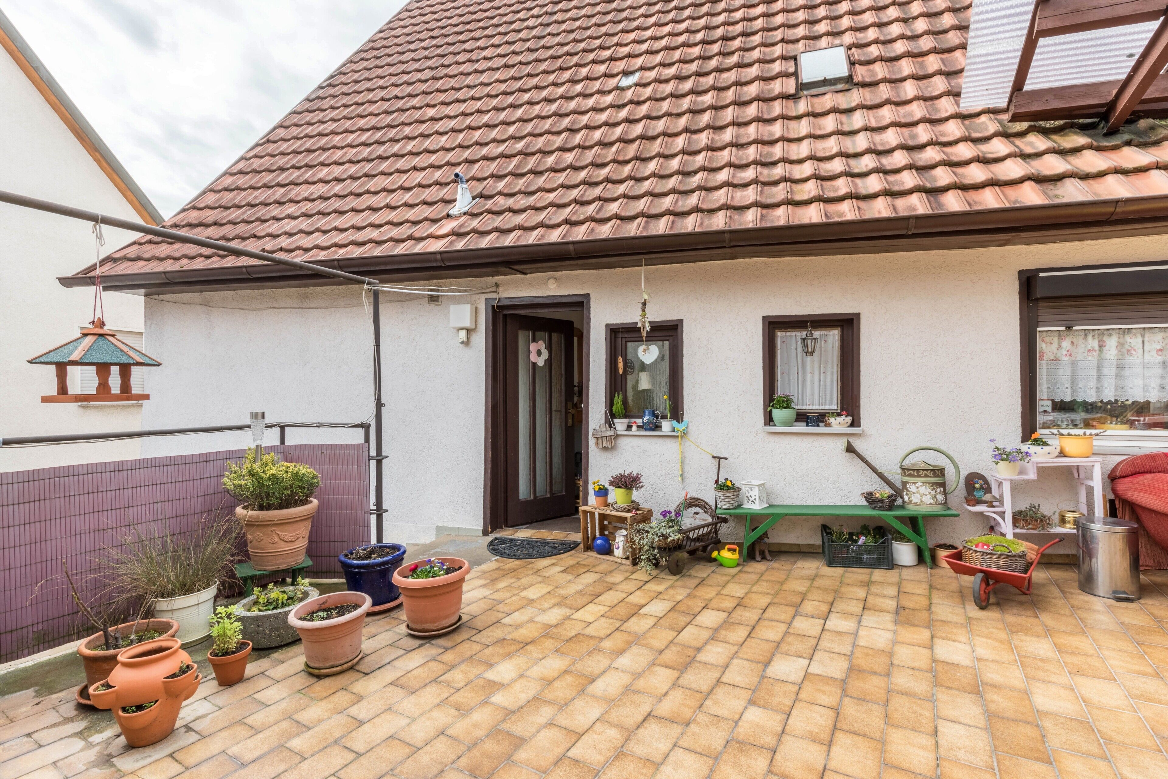 Una casa con tetto in tegole, una porta d'ingresso, una finestra e un balcone con vasi di piante e una carriola.
