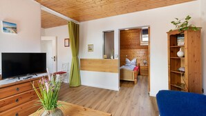Una habitación con techo de madera, un televisor sobre un aparador de madera, una cama con cabecera blanca, una silla azul y una estantería de madera.